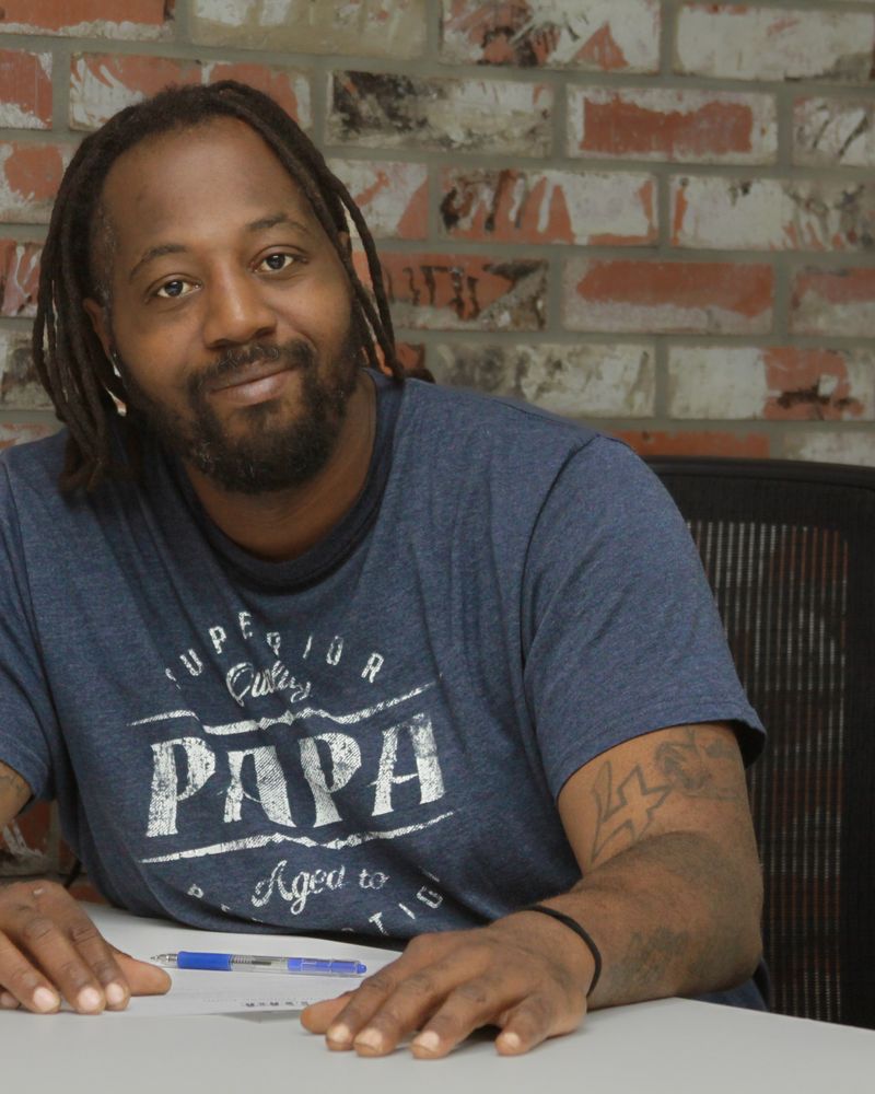 Marcus N. smiles while seated at a table in front of a brick wall.