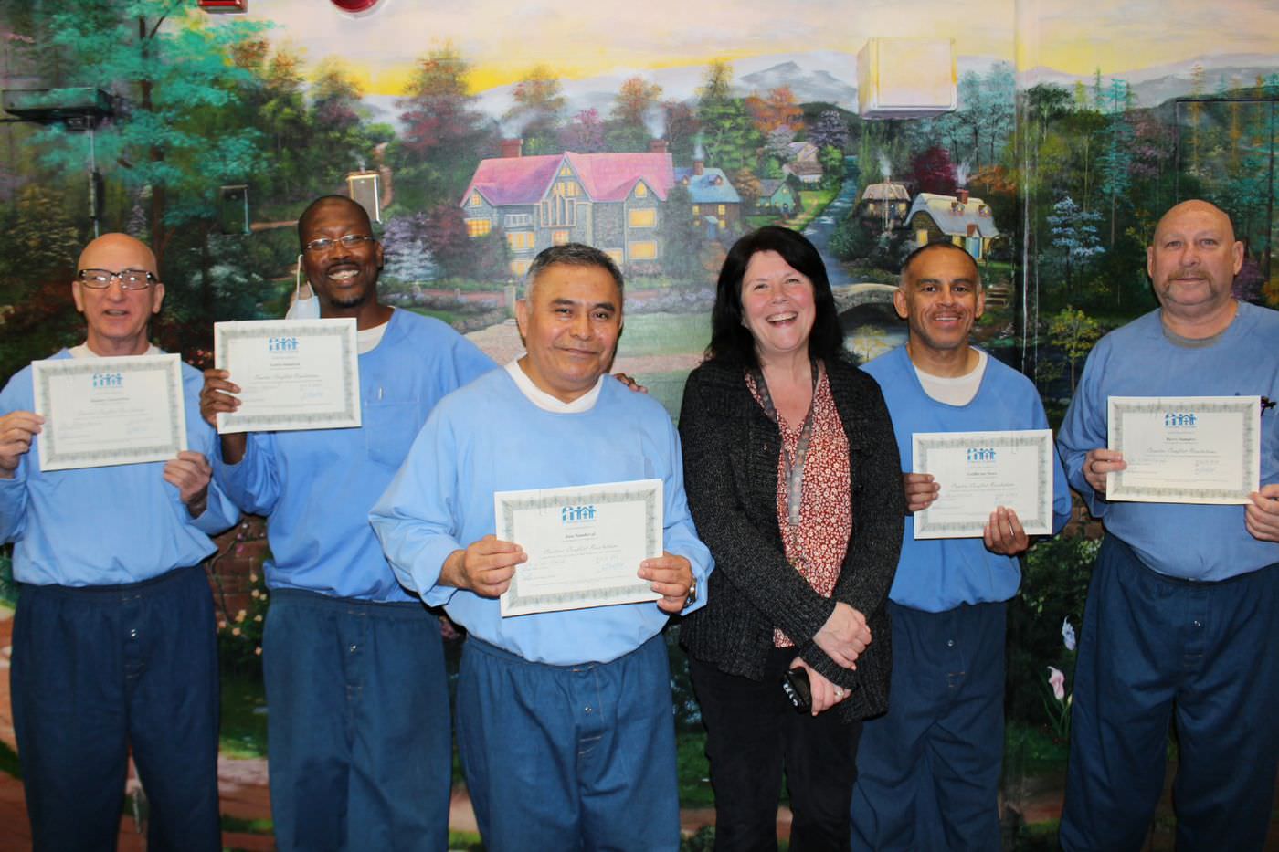 Five men in prison blues hold class certificates next to a Friends Outside staff member, in front of a painted mural.